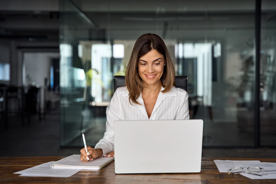 Une femme souriante travaille sur un ordinateur portable dans un bureau moderne avec cloisons vitrées